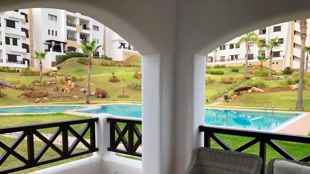 Un balcón con vista a una piscina y un jardín ajardinado, con un sofá de mimbre y plantas en macetas.