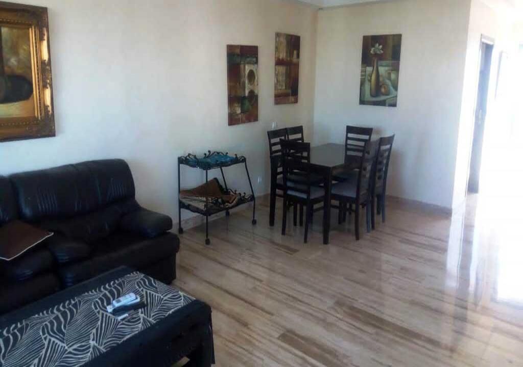 A living room with a black leather sofa, a patterned ottoman, a small table with a blue cloth, and a dining area with a wooden table and chairs.