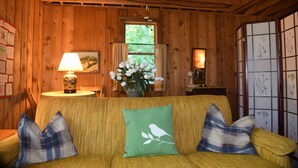 A cozy room with wooden walls, a sofa with plaid cushions, a table with a lamp, and a window with curtains. - The Violet Cottage (Frankfort)