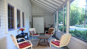A porch with white wooden walls, a wooden ceiling, and a view of a road and greenery. There are two white rocking chairs with red cushions and a small round table in the center. - The Violet Cottage (Frankfort)