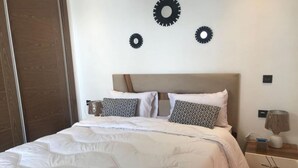 Una cama bien hecha con ropa blanca y almohadas estampadas, una mesita de noche de madera con una lámpara y decoración en la pared.