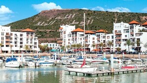 A marina with boats docked, surrounded by multi-story buildings with red roofs, and a hill in the background. - Apartment El Farah 10 min from the beach 4622 (Agadir)