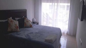 A neatly made bed with floral pillows, a bedside table with a lamp, and a view of the outdoors through sheer curtains. - Residence Adan Beach 4724 (Aourir)