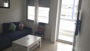 Una sala de estar con sofá, mesa de centro y una silla. Hay una ventana con persianas y un televisor montado en la pared.