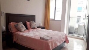 Un dormitorio con cama, almohadas, una mesita de noche y una ventana con cortinas.