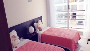 Een hotelkamer met twee bedden, elk met een roze sprei, een nachtkastje met een lamp en een raam met uitzicht op gebouwen.