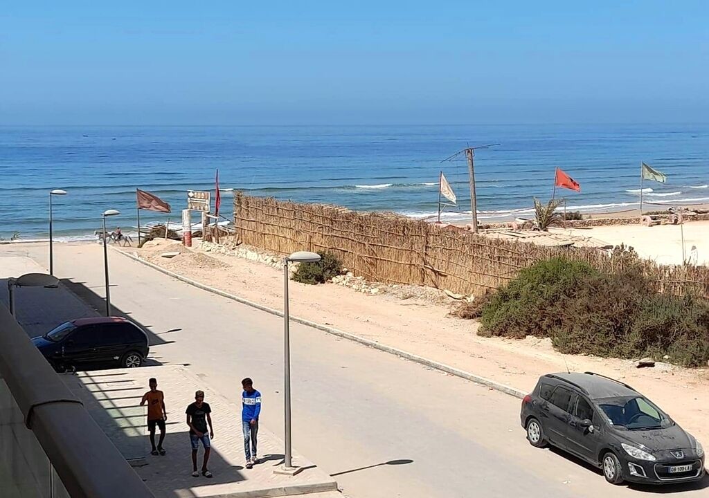 Una carretera costera con coches aparcados, un paseo peatonal y banderas cerca de la playa.