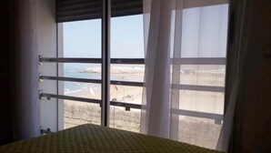 A room with a bed, a window with metal bars, and a view of a beach and rocky landscape. - Residence Adan Beach 4811 (Aourir)