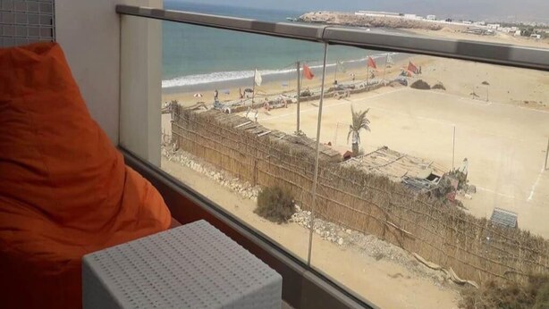Une chambre avec une grande fenêtre donnant sur une plage. On y trouve un lit avec un oreiller orange et un oreiller blanc sur une taie d’oreiller blanche.