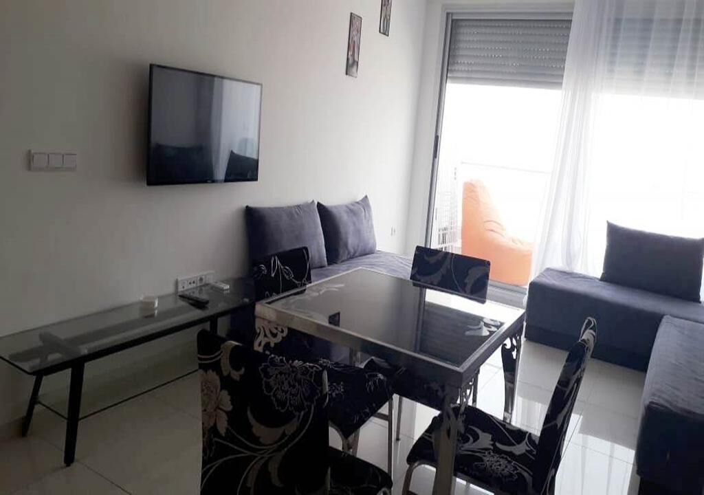 Un salon moderne avec une table à manger au plateau en verre, des chaises à motifs noir et blanc, un téléviseur à écran plat fixé au mur et un canapé avec des coussins.