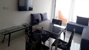 Un salon moderne avec une table à manger au plateau en verre, des chaises à motifs noir et blanc, un téléviseur à écran plat fixé au mur et un canapé avec des coussins.