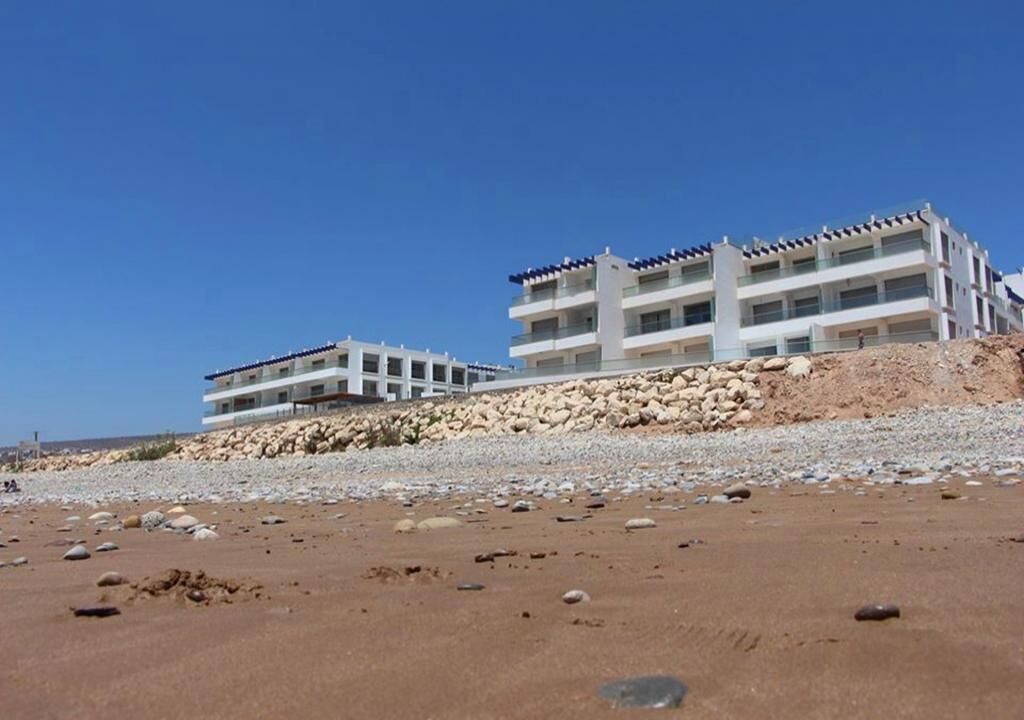 A coastal hotel with multiple floors, a rocky shoreline, and clear skies.