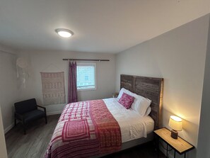 Ett sovrum med en säng, en stol, ett sängbord med en lampa, ett fönster med persienner och en väggdekoration.