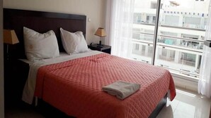 Een hotelkamer met een groot bed, twee nachtkastjes met lampen, een raam met uitzicht op gebouwen en een opgemaakt bed met een gevouwen handdoek.