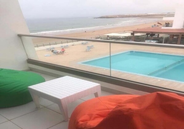 A balcony with a pool, beach view, and seating area.