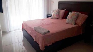 A neatly made bed with a pink bedspread and pillows, a dark headboard, and a nightstand with a lamp.