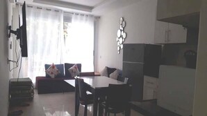 A modern living room with a sofa, dining table, and chairs. There is a kitchen area with a refrigerator and cabinets. - Residence Adan Beach 4797 (Aourir)
