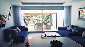 Una sala moderna con dos sofás, una mesa de centro y un ventanal corredero que da a un patio exterior con mobiliario de mimbre y vistas a un jardín.