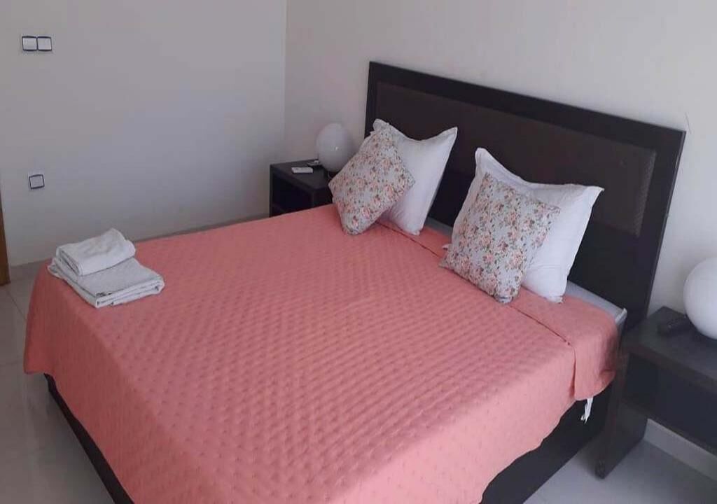 A neatly made bed with a pink bedspread, two pillows with floral patterns, and a folded white towel on the side.