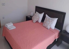A neatly made bed with a pink bedspread, two pillows with floral patterns, and a folded white towel on the side.