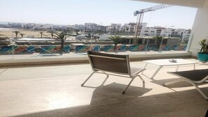 Un balcón con vistas a la playa y a una zona de construcción, con una silla de estructura metálica y una pequeña mesa.