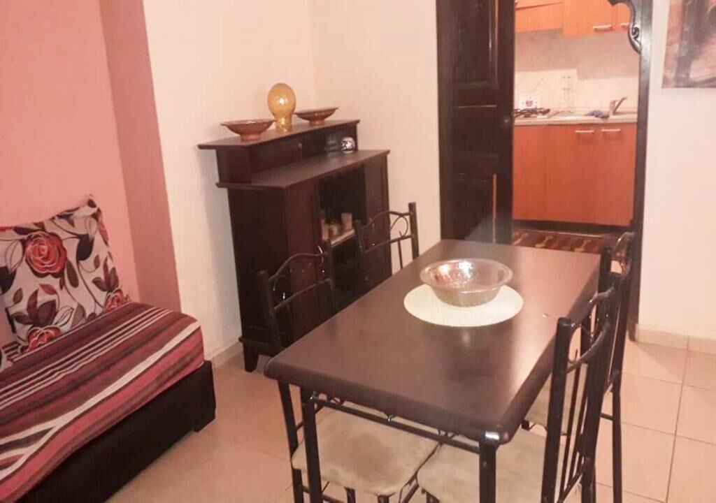 Ein kompakter Wohnbereich mit Esstisch und Stühlen, einer Couch, einem kleinen Schrank und einer durchgehenden Tür erkennbaren Küchenzeile.