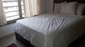 Una cama bien hecha con sábanas blancas en una habitación con cabecera de madera y vista al exterior a través de unas cortinas.