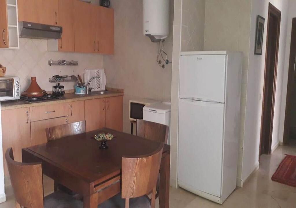 Een kleine keuken met houten kastjes, een witte koelkast, een fornuis en een eettafel met stoelen.