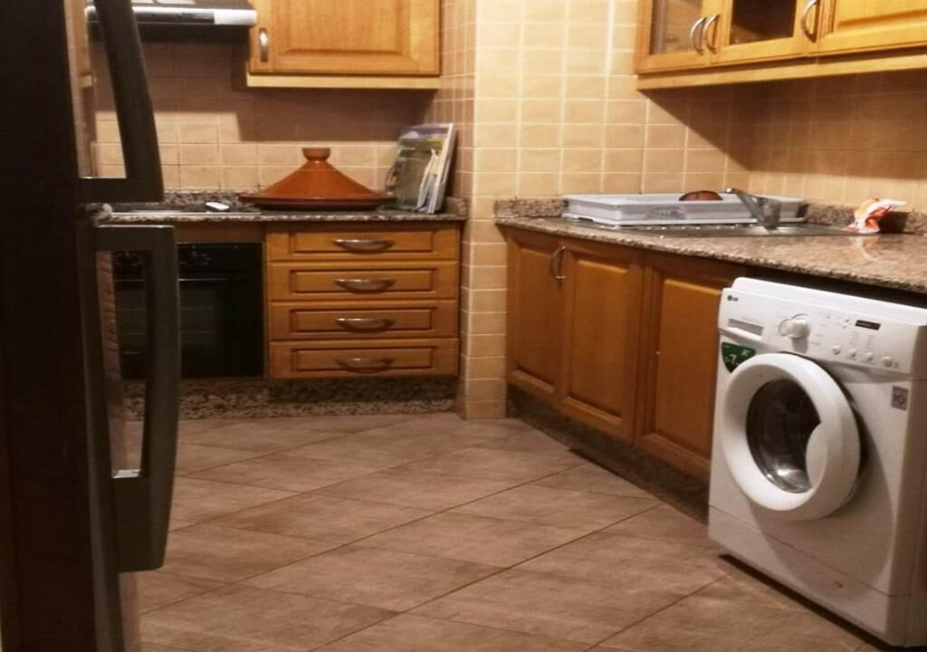 A compact kitchen with wooden cabinets, a built-in oven, a washing machine, and a countertop with various items.