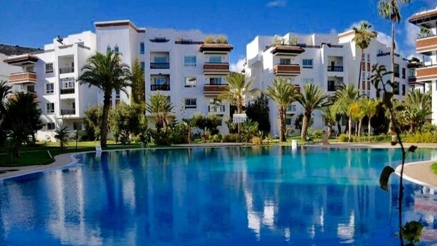 Une piscine entourée d’un bâtiment blanc doté de balcons, de palmiers et surplombée d’un ciel bleu clair.