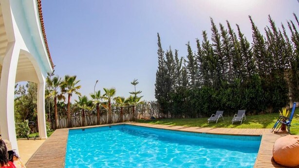 A swimming pool with a wooden deck, surrounded by palm trees and a fence. - Sublime Villa with swimming pool in Illigh Agadir 4906 (Agadir)