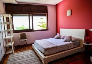 Ein Schlafzimmer mit Bett, Sessel, Ablagefläche und Fenster mit Blick auf Grünfläche.