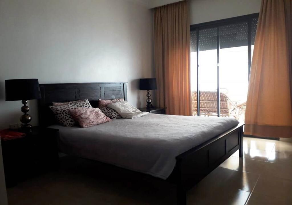 A neatly made bed with a headboard, two bedside tables with lamps, and a view of a balcony through large windows with curtains.