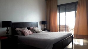 A neatly made bed with a headboard, two bedside tables with lamps, and a view of a balcony through large windows with curtains. - Holiday apartment in Résidence Fatima Zahra 4909 (Agadir)