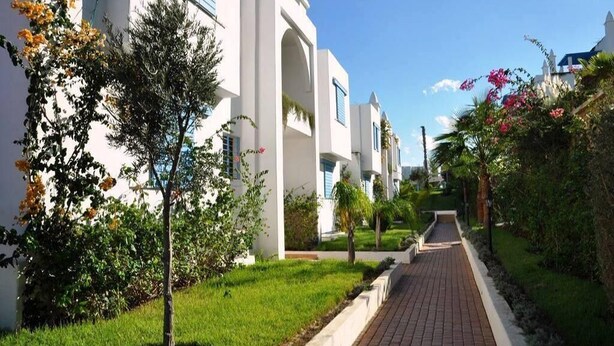 A pathway leading to a row of white buildings with arched windows, surrounded by greenery and flowering plants. - Residence Kabila Vista 4941 (Mdiq)