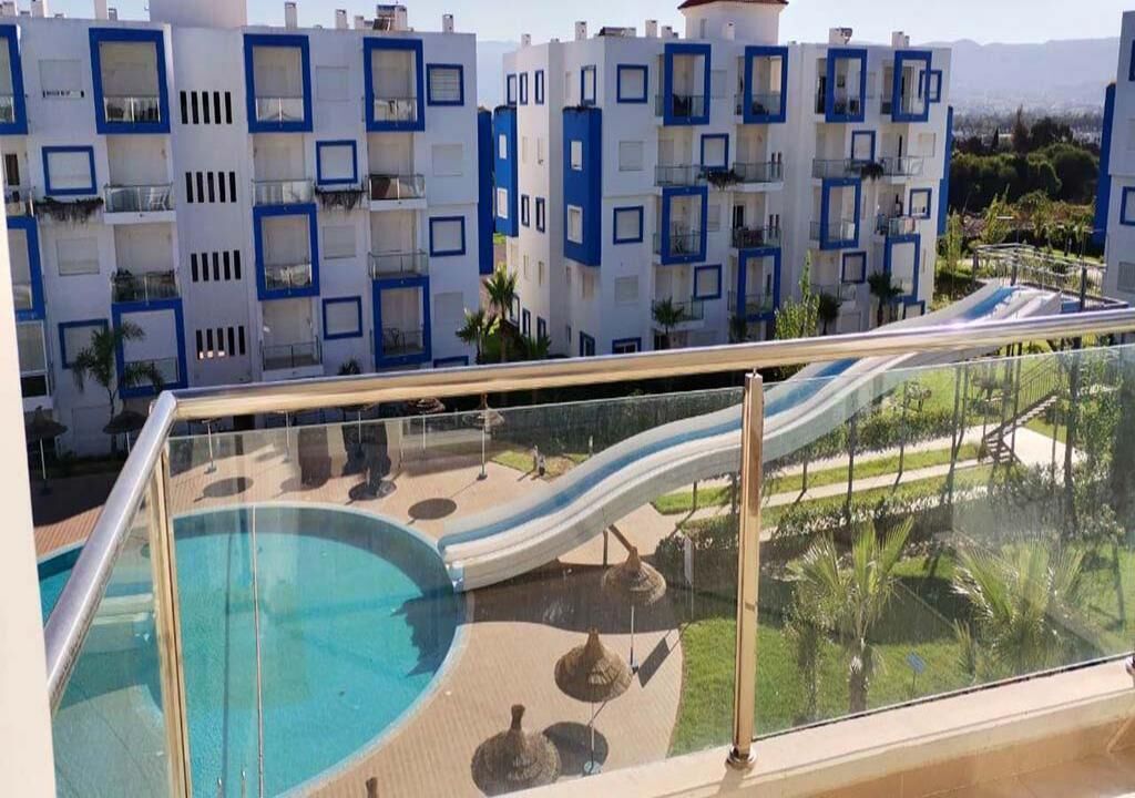 A pool area with a slide, surrounded by a glass railing, and buildings with balconies in the background.