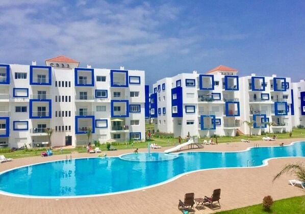 A modern hotel with multiple white and blue buildings, a large swimming pool, and outdoor seating areas.