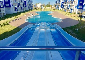 A water slide leading to a pool surrounded by modern buildings with balconies. - Residence Kariat Cabo 4951 (Cabo Negro)