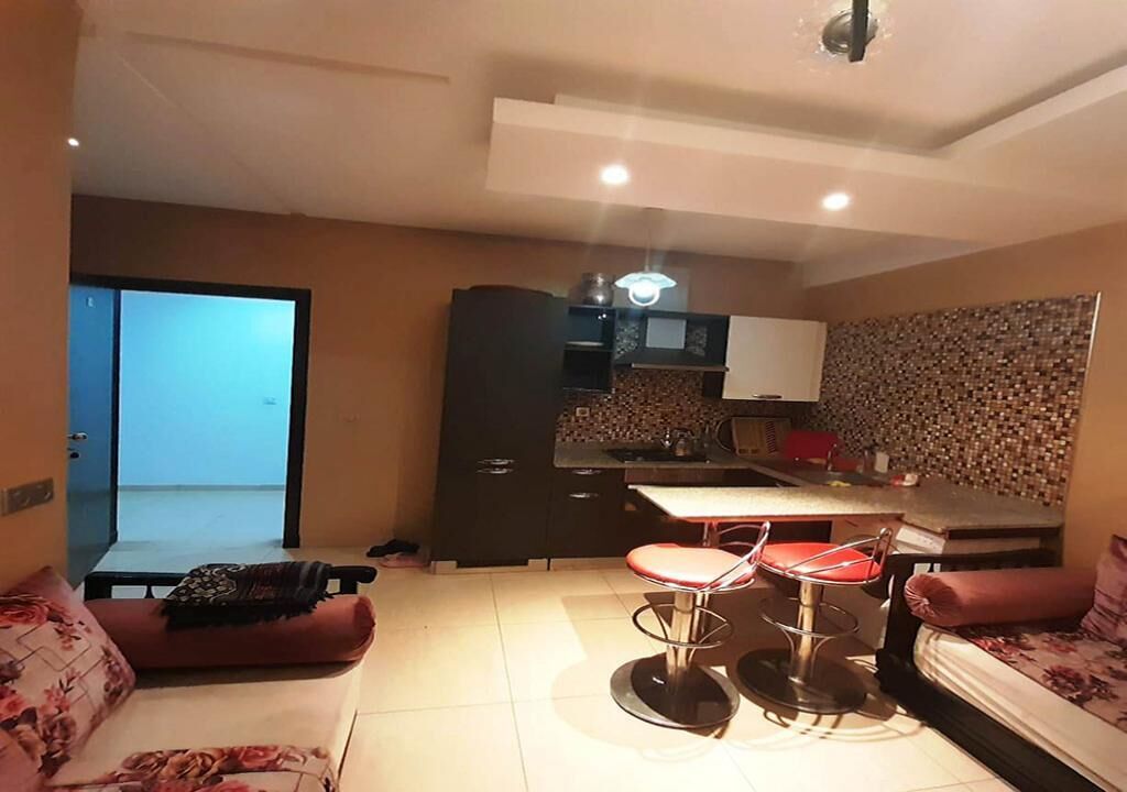 Een knus woongedeelte met een kitchenette, twee rode barkrukken, een bank en een deur naar een balkon.