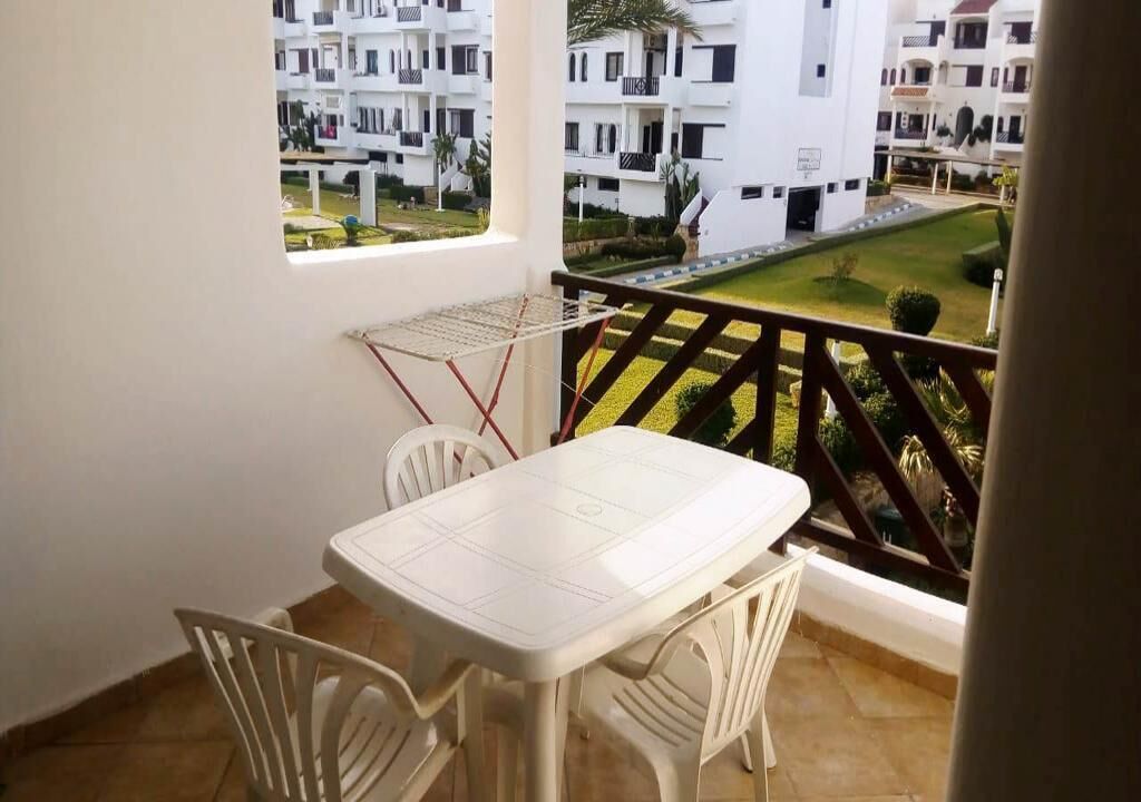 A balcony with a white table and chairs, overlooking a well-maintained garden and residential buildings.