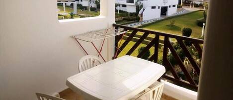 A balcony with a white table and chairs, overlooking a well-maintained garden and residential buildings.