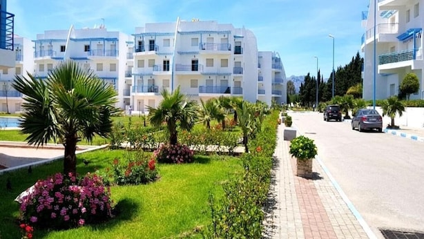 A modern apartment building with multiple balconies, a well-maintained garden with palm trees and flowers, and a clear blue sky. - Residence Riviera Beach 5024 (Cabo Negro)