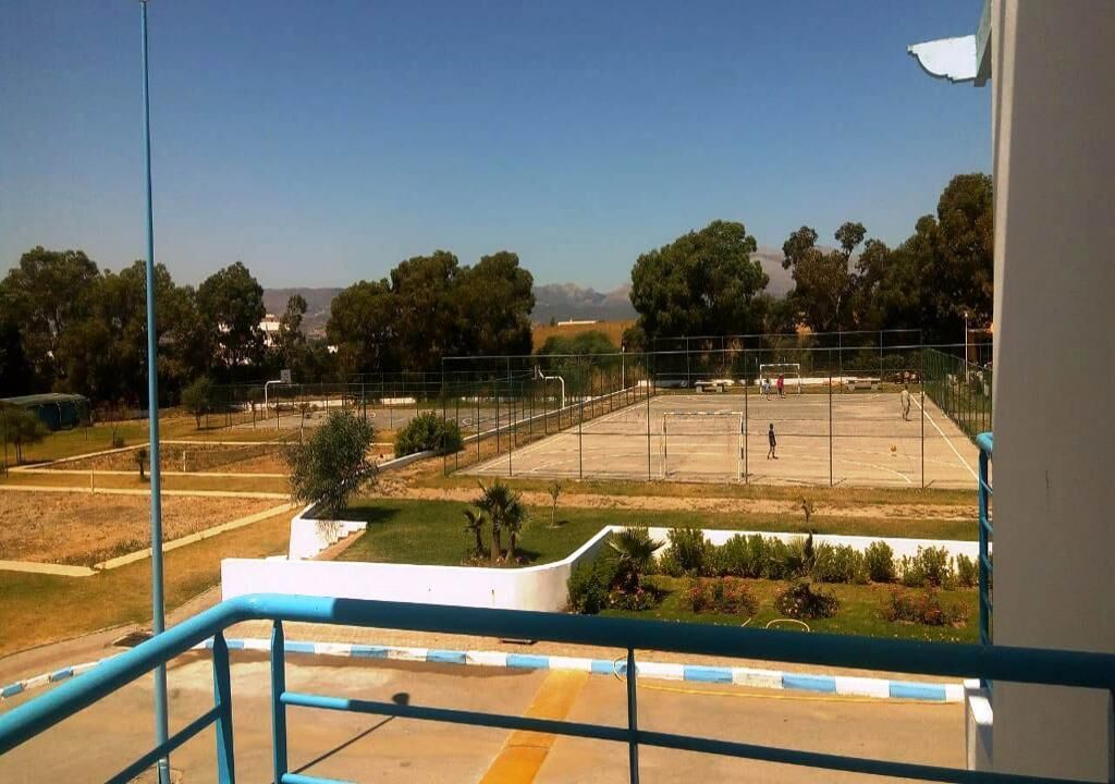 Une vue depuis un balcon donnant sur un terrain de sport, avec un ciel dégagé et des arbres à l’horizon.
