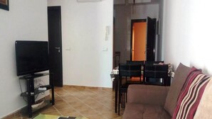 Un salon comprenant un canapé, un téléviseur posé sur un meuble, une table à manger avec des chaises, et une porte donnant sur une autre pièce.