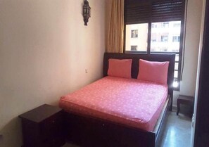 A single bed with a wooden frame and a pink mattress in a room with a window and a wall-mounted light fixture.
