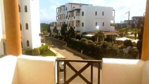 A balcony with a view of a multi-story building with balconies, a street, and some greenery.
