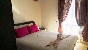 Un lit bien fait, avec un drap blanc et des oreillers roses, une serviette pliée posée sur le lit, et une table de chevet avec une lampe.