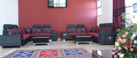 A lobby with a red wall, a flat-screen TV mounted above a sofa set, and a patterned rug on the floor.