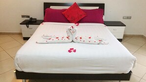 Uma cama bem arrumada com um cobreleito branco, duas toalhas brancas dispostas em formato de coração, e um único pétalo de flor vermelha sobre a cama.