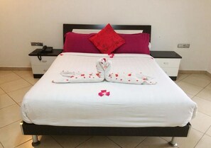 A neatly made bed with a white bedspread, two towels arranged in a heart shape, and a single red rose petal on the bed.